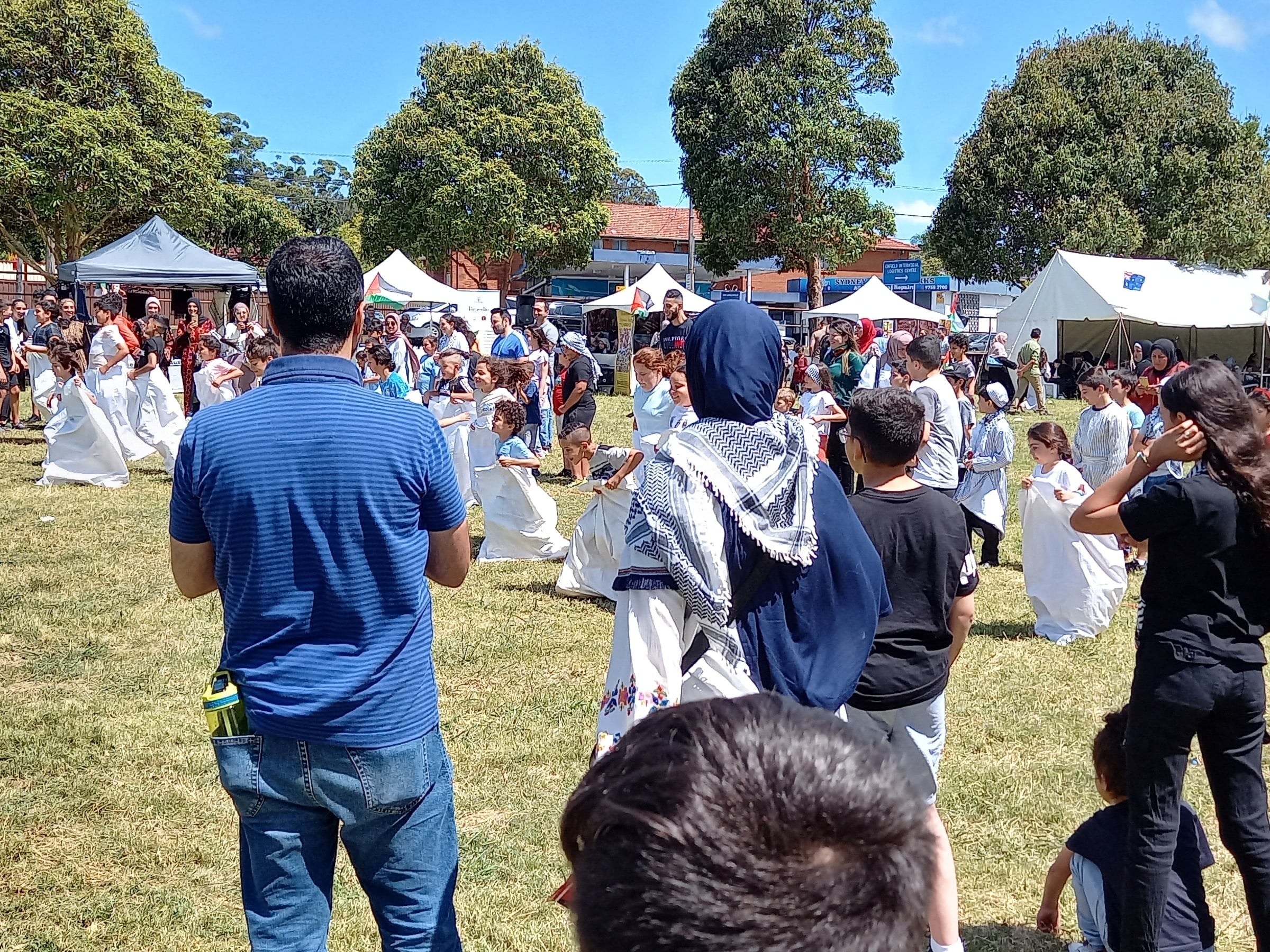 Families cheering on children in the sack race, a woman in a keffiyeh scarf in the middle